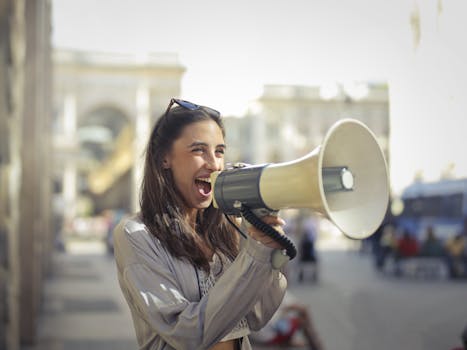 PPRA / IREM Collaboration Cheerful young woman in a casual outfit shouting into a megaphone on a sunny day.